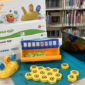 Photo of the Plugo STEM Kit contents on a gray table: four boxes, yellow tiles, an orange plastic tile holder, a yellow plastic figure, a teal table top pad, and a black tablet.