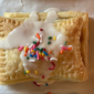 Photo of a finished "Cutie Tart" rectangle pastry, with white icing and multi-colored sprinkles, on a parchment lined cookie sheet.