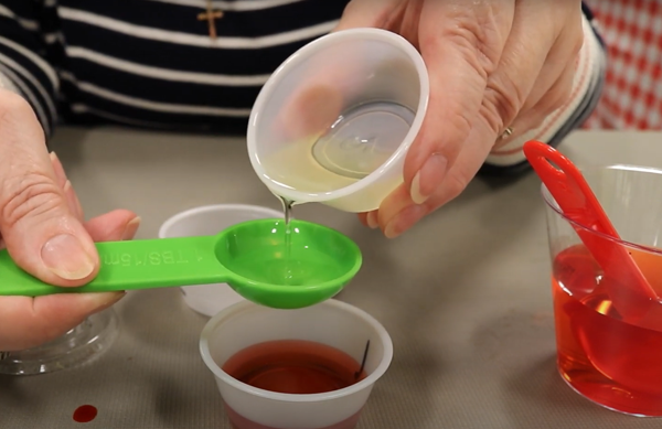 Photo of a woman pouring oil from a small cup into a green tablespoon. She is holding the tablespoon over another small cup with a red liquid in it.