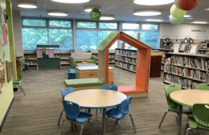 Play Spot area with two small tables in the foreground, a tall house-like open reading nook, activity tables, and a couch in the back for caregivers.